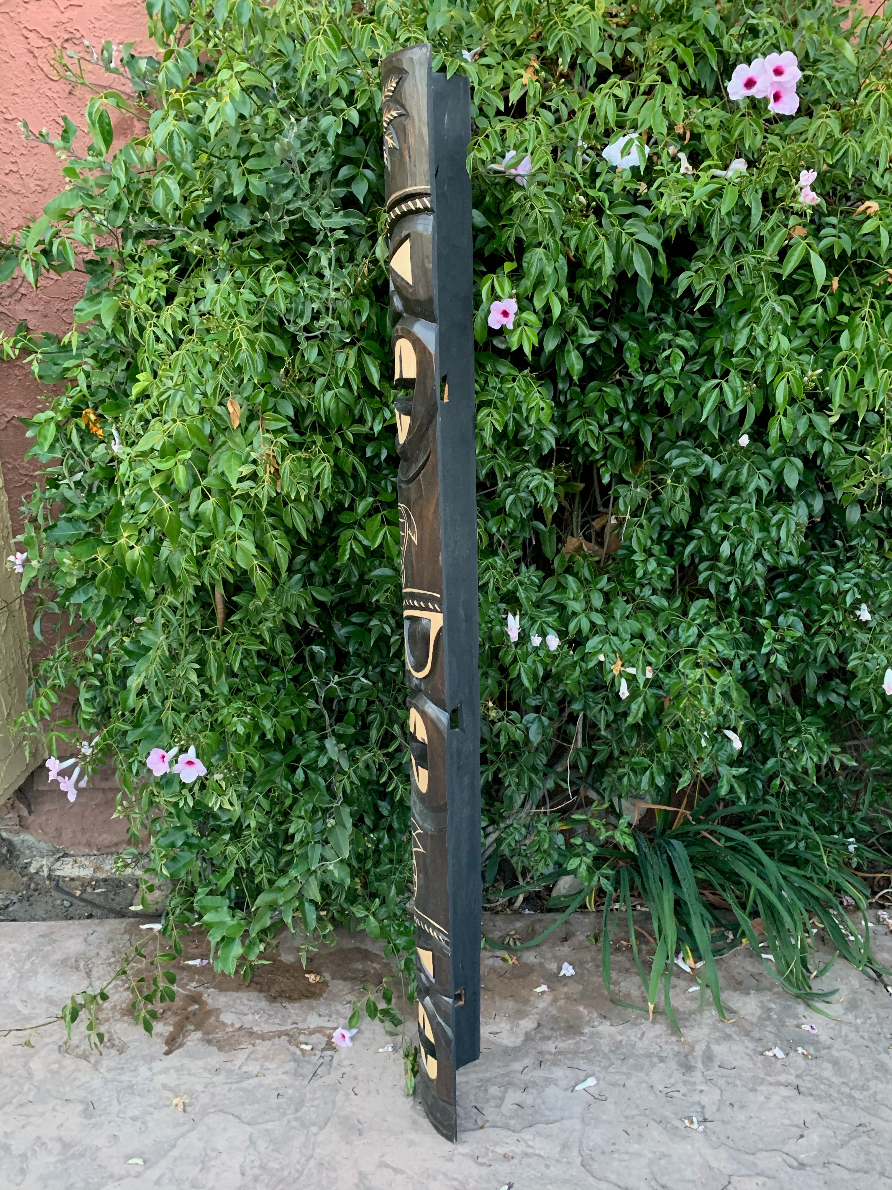 Dark wooden totem-style sculpture leaning against a leafy shrub with pink hibiscus flowers