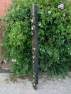 Dark wooden totem-style sculpture leaning against a leafy shrub with pink hibiscus flowers