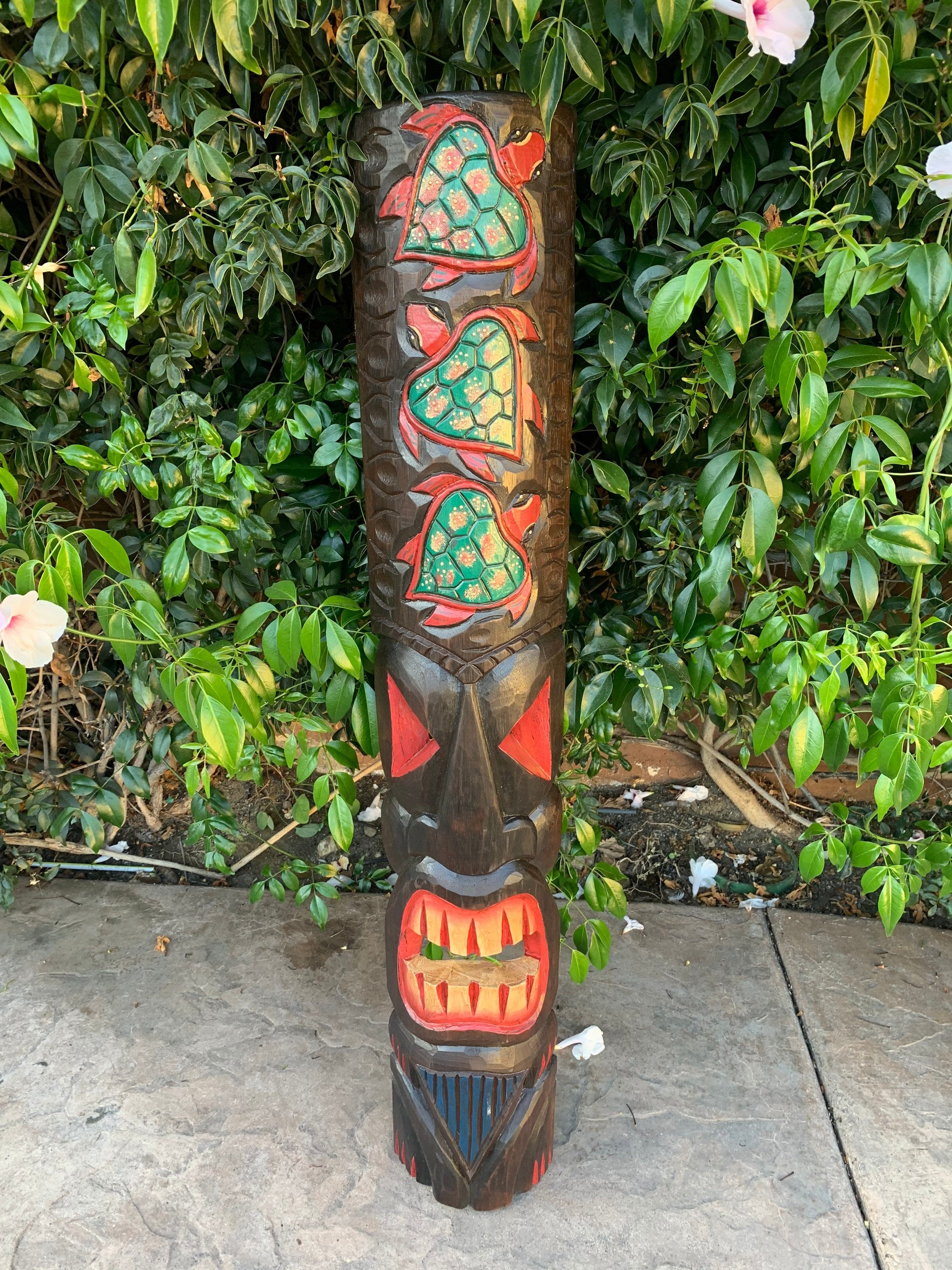Wooden totem pole with green turtle carvings and a large red open-mouth face among greenery.