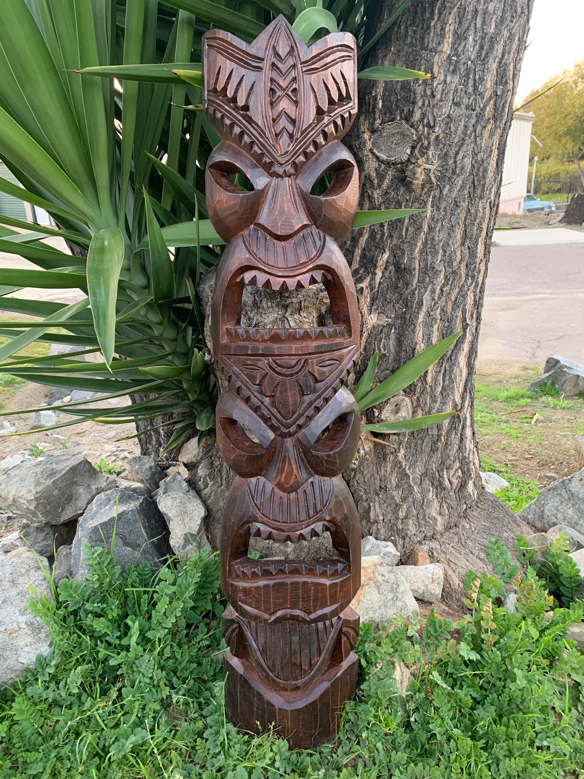 Stacked carved wooden totem pole with multiple faces beside a tree in a garden