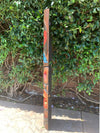 Carved wooden totem pole with orange, red, and blue geometric designs outdoors beside a green hedge