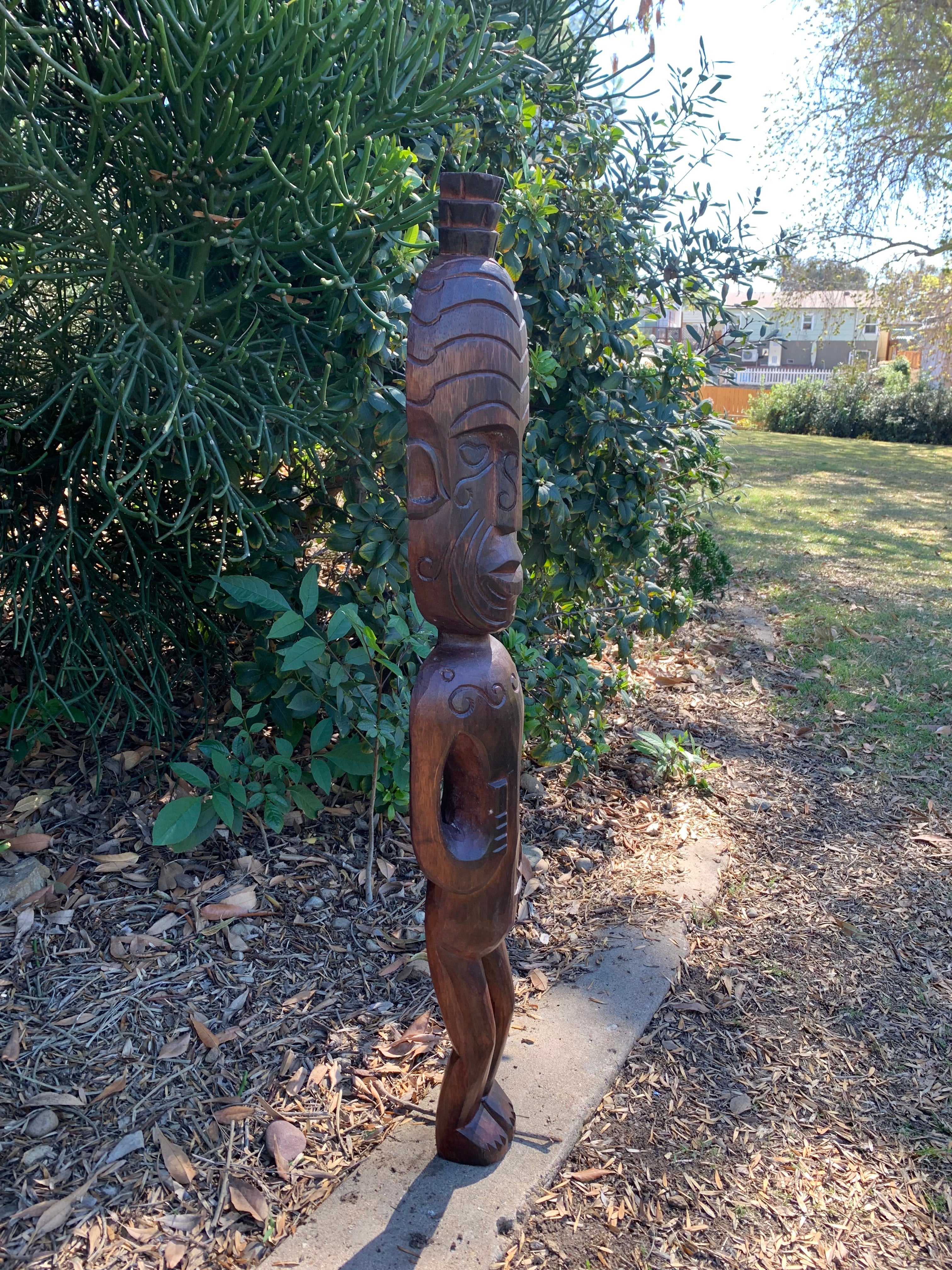 Carved wooden tribal statue standing outdoors beside a hedge in a garden.