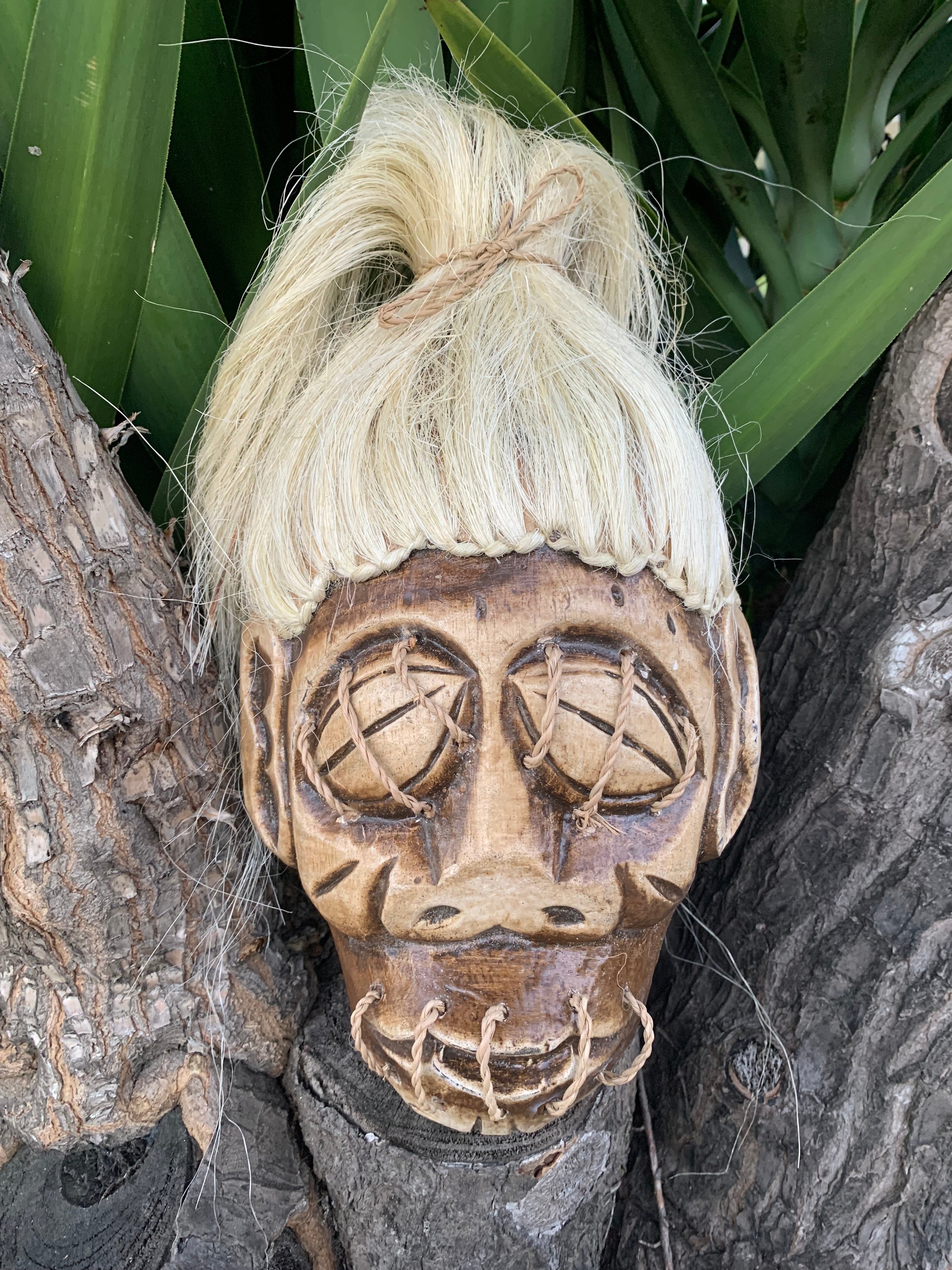 Wooden tribal mask with a blonde wig and rope-laced eyes on a tree trunk
