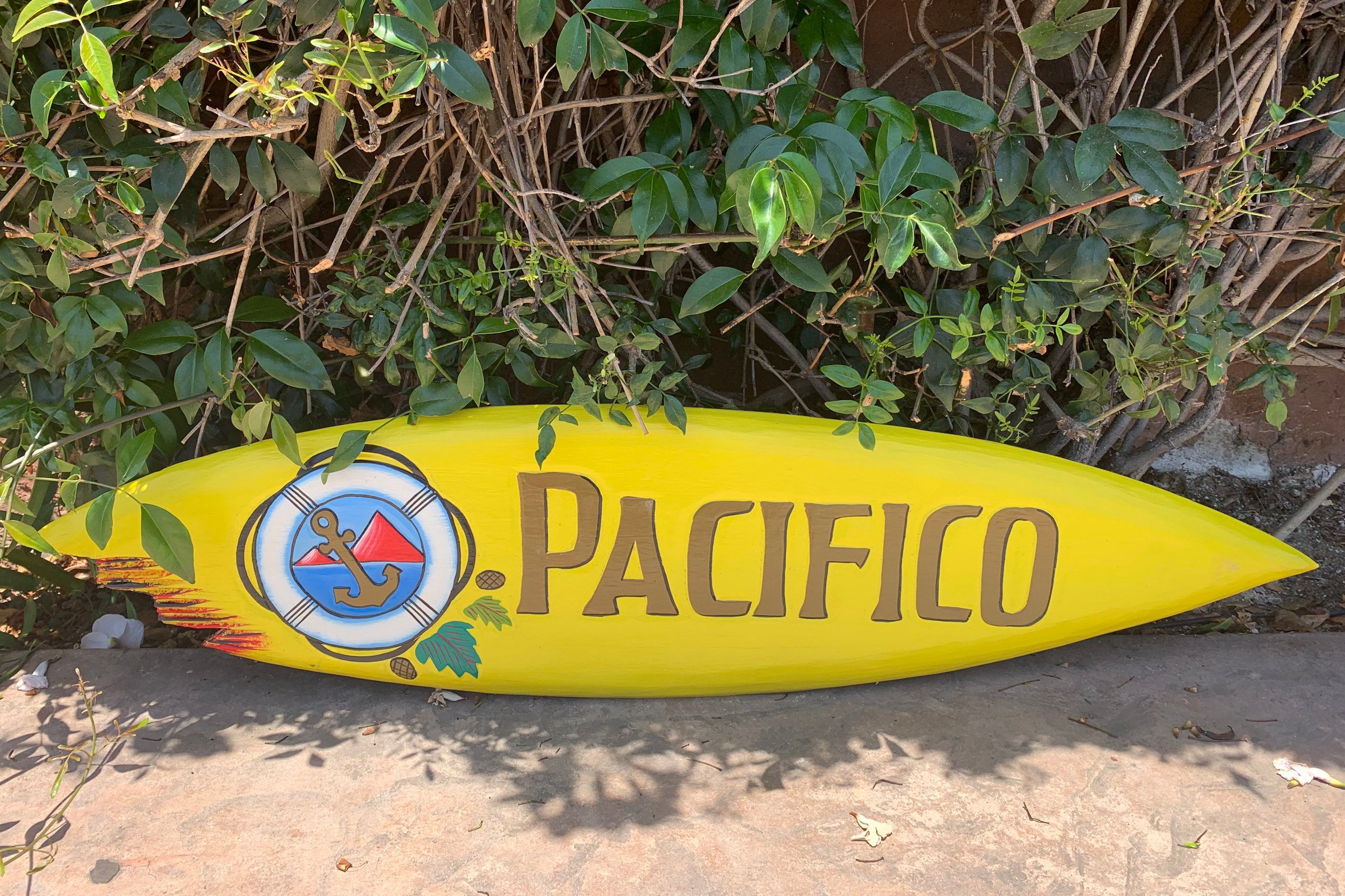 Yellow Pacifico surfboard with anchor logo and text resting on a sidewalk beside green foliage